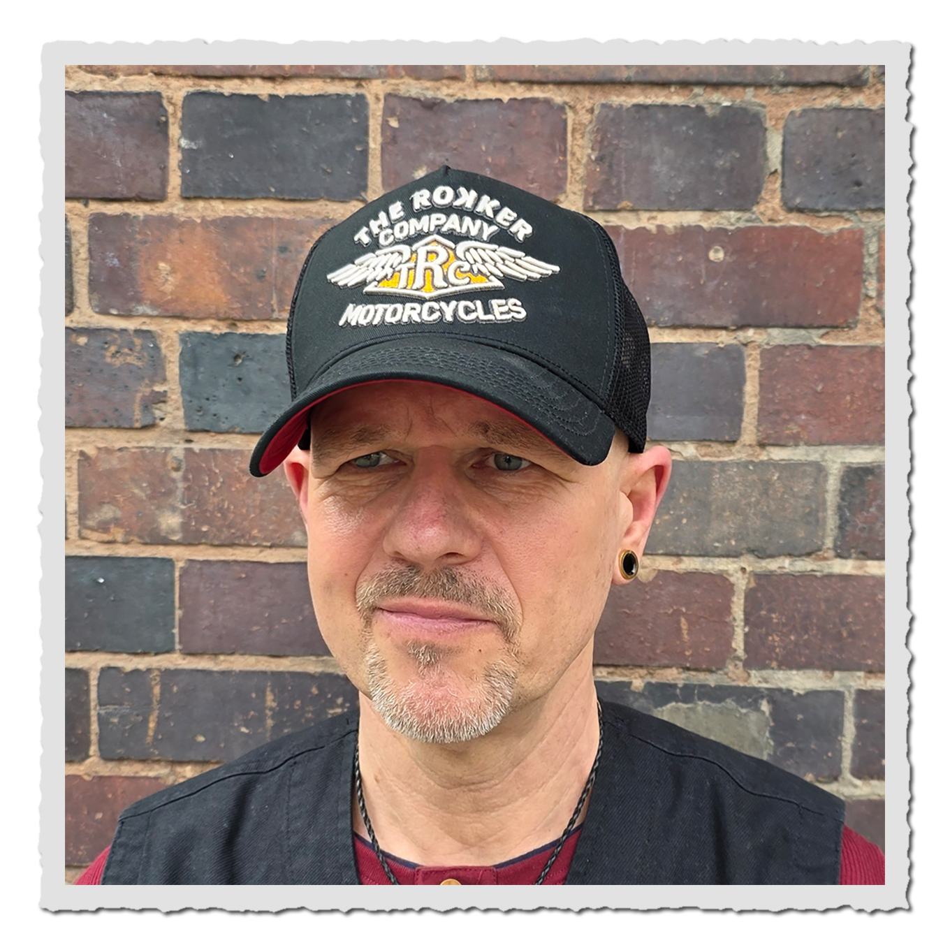 Porträt einer Person mit schwarzer Trucker Cap vor Ziegelwand, Cap-Logo frontal sichtbar, Tageslichtaufnahme.